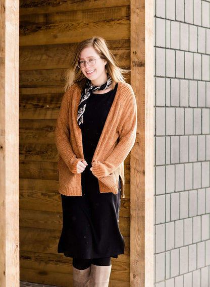 Woman wearing an open front cardigan that is chenille and camel in color. She is also wearing it over a black dress and brown riding boots. She is standing on the stairs at Inherit Clothing Company.