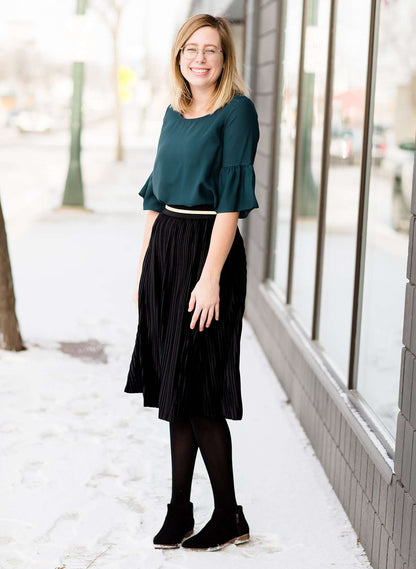 Woman wearing a black, velvet pleated midi skirt. This skirt has a gold accent around the elastic waistband. This modest skirt is paired with a teal, ruffle sleeve poets blouse.