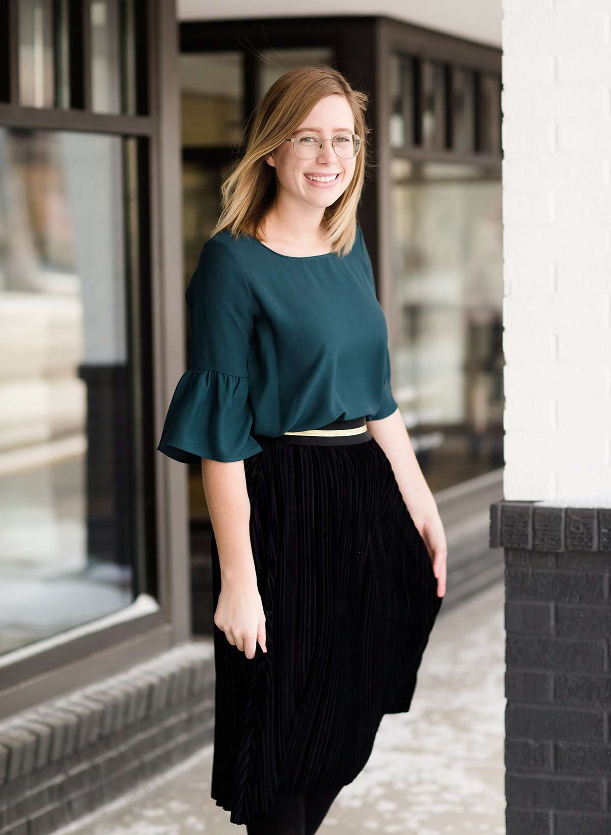 Woman wearing a black, velvet pleated midi skirt. This skirt has a gold accent around the elastic waistband. This modest skirt is paired with a teal, ruffle sleeve poets blouse.