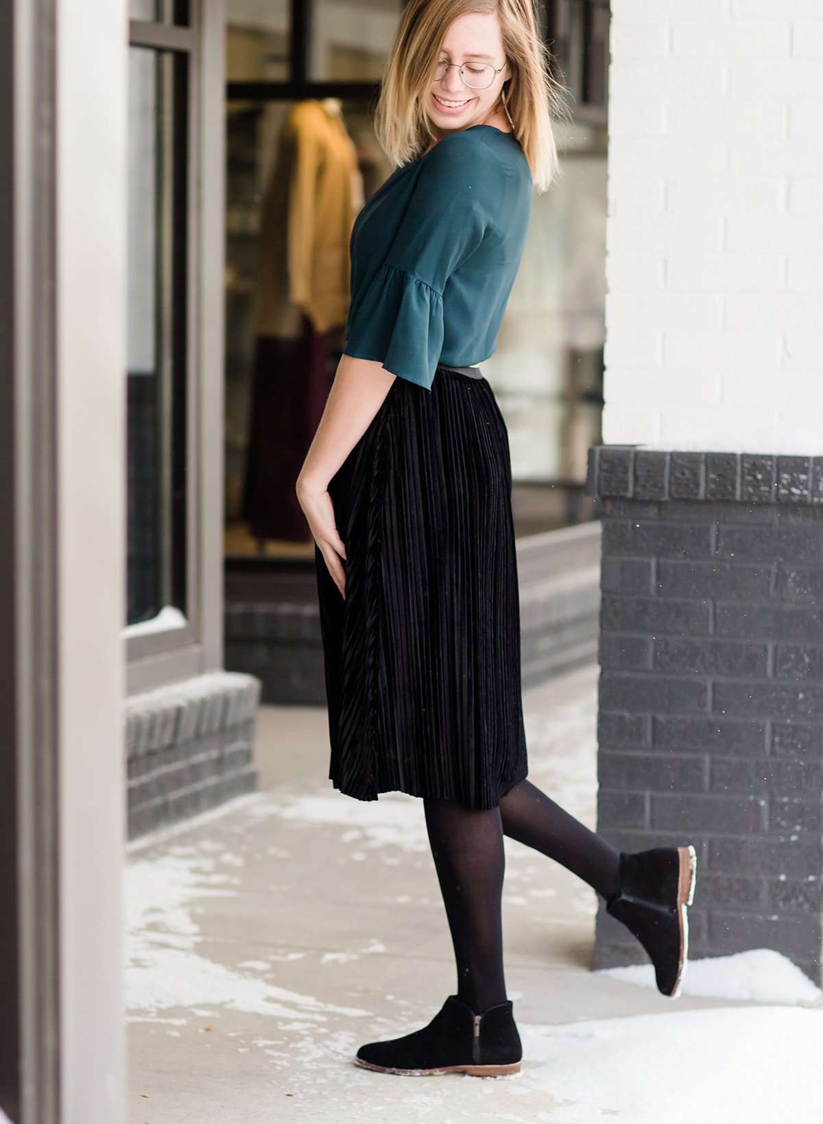 Woman wearing a black, velvet pleated midi skirt. This skirt has a gold accent around the elastic waistband. This modest skirt is paired with a teal, ruffle sleeve poets blouse.