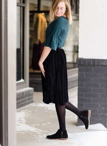 Woman wearing a black, velvet pleated midi skirt. This skirt has a gold accent around the elastic waistband. This modest skirt is paired with a teal, ruffle sleeve poets blouse.